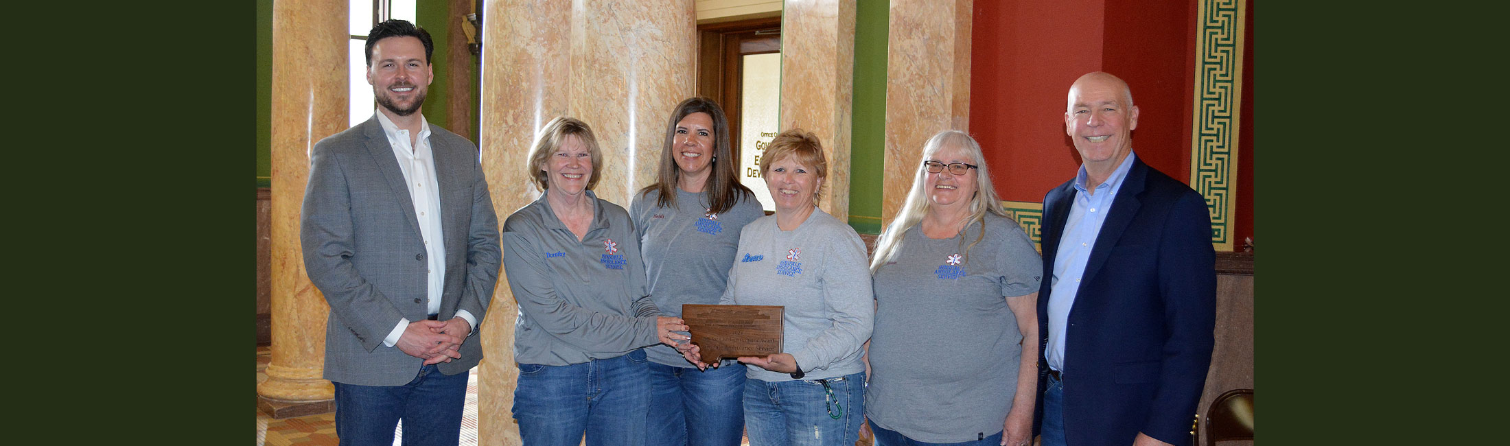 Governor Greg Gianforte and Director Charlie Brereton standing with Montana Emergency Medical Services (EMS) personnel holding award