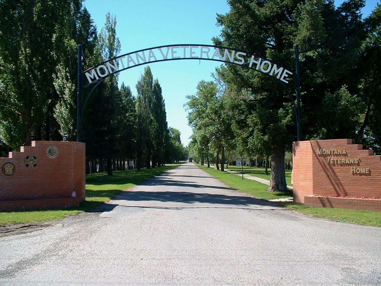 A picture of the gate outside of the Montana Veterans home. There is a road, an arch that says Montana Veterans Home, and trees.