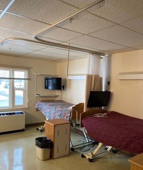 A picture of a resident room. There are two beds, with TVs above each bed. There is a window with an air conditioner below it, a night stand by the bed, and a trash can.