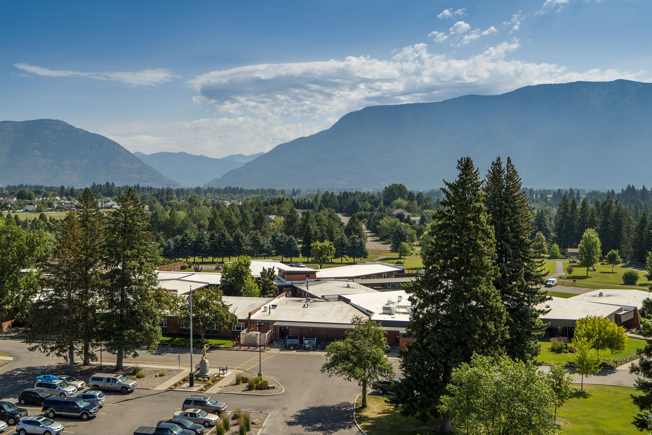 An aerial view of the grounds of the Montana Veterans home. There are large mountains in the background, as well as many trees. There is also a parking lot and several buildings.
