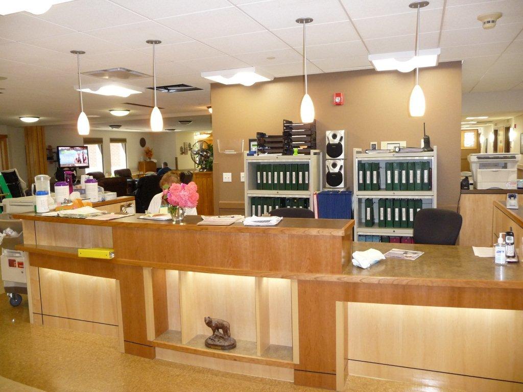 A picture of the west nurse’s station. There is a long half wall with chairs behind it for the nurses to sit on. There are binders on shelves and papers on the table.